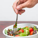 A hand holding a Reserve by Libbey Baroque stainless steel salad fork in a bowl of salad.