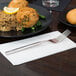 A Libbey stainless steel dinner fork on a white napkin next to a plate of food.
