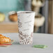 A close-up of a Choice double wall paper hot cup with a bean print filled with coffee on a table with a croissant.