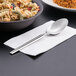 A Libbey stainless steel serving spoon on a napkin next to a bowl of pasta with sauce.