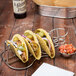 A Clipper Mill stainless steel guitar shaped taco holder with three tacos on a table.