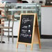 A natural oak Choice A-Frame sidewalk chalkboard with white writing on it in front of a bakery display.