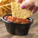 A person holding a chip and dipping it into a bowl of salsa on a table with a Carlisle black melamine ramekin of salsa.
