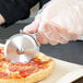 A person in a plastic glove using a Dexter-Russell pizza cutter to cut a pizza.