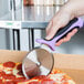 A hand with a purple and black Mercer Culinary pizza cutter cutting a pizza.