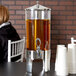 A woman sitting at a table with a Vollrath beverage dispenser filled with drinks.