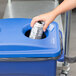 A hand puts a can of soda into a blue Rubbermaid recycling bin.