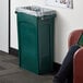 A woman sitting next to a green Rubbermaid Slim Jim trash can with a plastic bag over it.