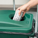 A hand putting a paper into a green Rubbermaid Slim Jim recycling container.