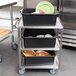 A Lakeside stainless steel utility cart with black containers holding white and green plates.