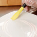 A hand using a yellow and black Mercer Culinary saw tooth silicone brush to plate a white dish.