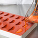 A person pouring orange liquid from a glass container into a red silicone tangerine slice mold on a counter.
