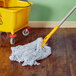 A Rubbermaid wet mop with a blue and white mop head in a room with a wooden floor and a yellow bucket.