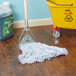 A Rubbermaid Web Foot Looped End Wet Mop on a wooden floor next to a yellow bucket.