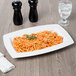 A rectangular white porcelain bowl filled with spaghetti on a table with a glass of water.