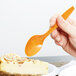A hand holding a Creative Converting pumpkin spice orange plastic spoon over a piece of cake.