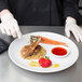 A person in gloves using Mercer Culinary precision plating tongs to cut a meal on a plate.