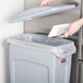 A person putting a paper into a Rubbermaid Slim Jim trash can with a grey lid.
