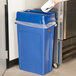 A hand putting a plastic bag into a blue Rubbermaid Slim Jim trash can with a blue lid.