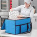 A woman in a kitchen using a blue Choice insulated food delivery bag to carry a baking tray.