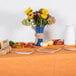 A table with a Creative Converting Pumpkin Spice Orange table cover, flowers in a blue vase, and plates of food.