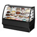 A True black refrigerated curved glass bakery display case on a counter with cakes inside.
