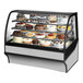 A True refrigerated bakery display case with cakes and desserts behind curved glass.