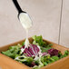 A black Carlisle salad spoon pouring dressing over a salad.