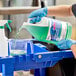 A person in blue gloves pouring Noble Chemical Reflect Super Concentrated Window Cleaner into a blue container.