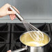 A hand using a Vollrath stainless steel whisk to stir a pot of liquid.