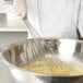 A person in a white chef's uniform using a Vollrath stainless steel French whisk to whisk yellow liquid in a bowl.