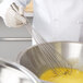 A person in white gloves using a Vollrath stainless steel whisk to mix eggs in a bowl.