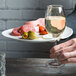 A person holding a Tuxton bright white china appetizer plate with a glass of wine and food.