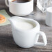 A Tuxton bright white china creamer filled with milk on a white surface next to a cup of coffee.