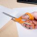 A hand with orange and gray Fiskars scissors cutting a piece of paper.