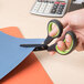 A person using Universal carbon-coated industrial scissors with black and green bent handles to cut a piece of paper.