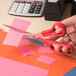 A person using Universal stainless steel scissors with a red bent handle to cut pink paper.