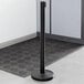 A Lancaster Table & Seating black metal crowd control stanchion with a retractable belt.