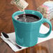 A turquoise Fiesta china mug on a napkin with liquid being poured into it using a spoon.