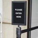 A white sign that says "Please Enter Here" in a Lancaster Table & Seating black vertical stanchion sign frame.
