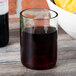 A clear Arcoroc wine tumbler filled with red wine on a table in front of a bowl of fruit.