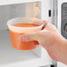 A hand holding a ChoiceHD translucent plastic deli container of soup in front of a microwave.
