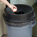 A person's hand putting an apple in a gray and black round trash can with a funnel top lid.