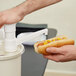 A person using a white Cambro condiment dispenser fixed nozzle pump to add mustard to a hot dog.