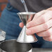A person using an American Metalcraft stainless steel jigger to pour liquid into a metal cup.
