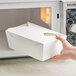 A hand holding a white Choice paper take-out box of food in front of a microwave.