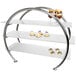 A stainless steel 3 tier circular display stand with glass shelves holding desserts.