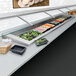 A counter with a Hatco slanted drop-in cold food well holding black plastic containers of food.