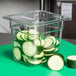A Cambro clear polycarbonate food pan with sliced cucumbers in it on a green surface.