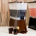 A Tablecraft brown beverage dispenser with dark liquid and two glasses of liquid.